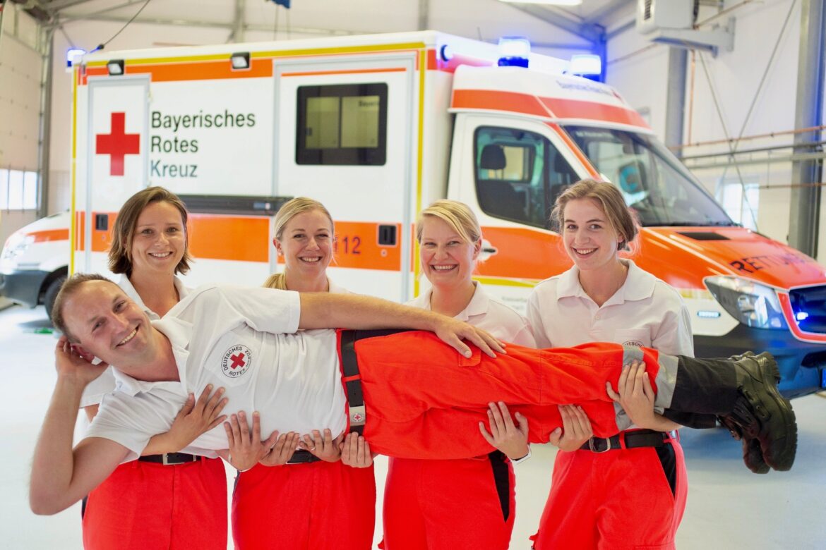 Rettungskräfte des BRK stehen vor einem Rettungsauto. Vier Damen Tragen einen Heeren. Alle fünf Personen sehen Glücklich aus